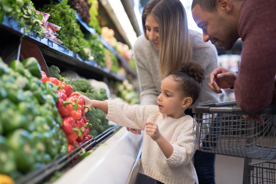 A child chooses a pepper from the fruit and vegetable section of a grocery store. The parents stand next to them. 