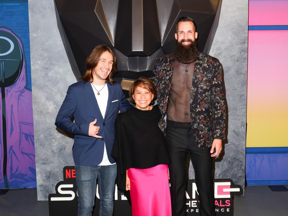 phill, mai, and sam out of the game in casual party wear at a squid game party, with their arms around each other and smiling