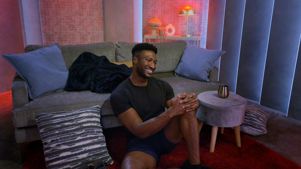 Uche sits in the floor of the pods in Netflix's "Love Is Blind" season 5, with his back against a couch covered in pillows and blankets. He's smiling, wearing a black shirt and shorts, and a tumbler of wine is perched on the ottoman next to him. 