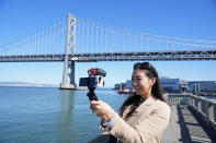 Content creator Cynthia Huang Wang works below the San Francisco-Oakland Bay Bridge in San Francisco, Monday, April 8, 2024. Despite a strong job market, there are still thousands of people who have found themselves out of work across industries stretching from tech to retail to media. But rather than trying to find another job in their old role, some workers are turning to online content creation. (AP Photo/Eric Risberg)