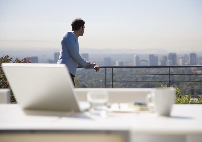 A man looking out at a city on a rooftop with a table and laptop in the foreground.