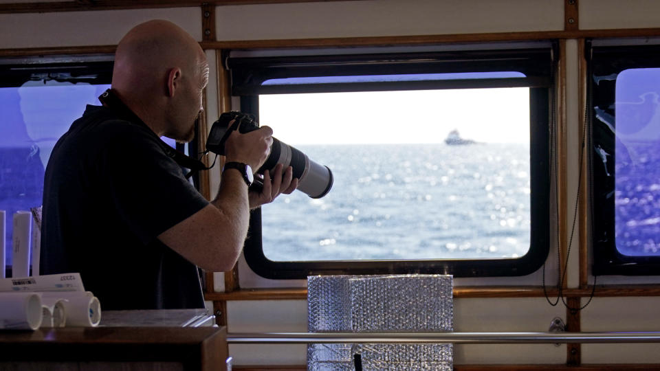 Engineer John Rayfield, of Mobile, Ala., photographs foreign ships and crews installing portions of a wind farm, Tuesday, July 11, 2023, off the coast of Rhode Island. The trade association that represents the offshore service industry is going to great lengths to make sure that jobs go to Americans as the U.S. offshore wind industry ramps up. (AP Photo/Charles Krupa)