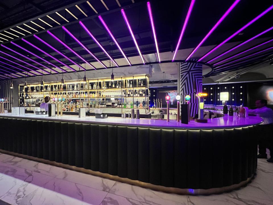 The main bar at The NinetyThird in The O2 includes beers on tap. One bartender is visible and lots of drinks on the shelves behind, and purple LED lights on the ceiling.