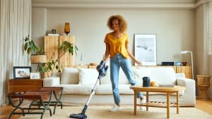 Smiling young woman cleaning carpet with vacuum cleaner at home. Female is doing housework in living room. She is standing against sofa.