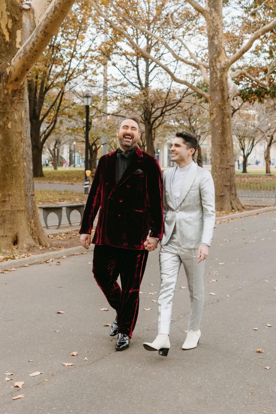 Two grooms hold hands and walk through a park.