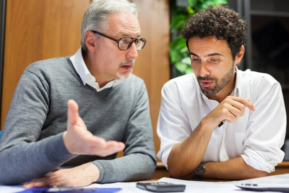 Older man and younger man have a discussion as a calculator sits in front of them on the table.