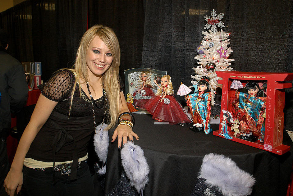 Smiling Hilary leaning on a table with several Bratz dolls on it