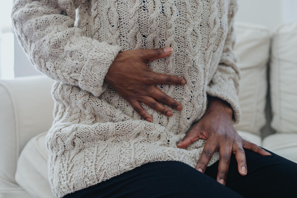 A person sitting on a couch holds their stomach and side, appearing to have discomfort, while wearing a knitted sweater and dark pants