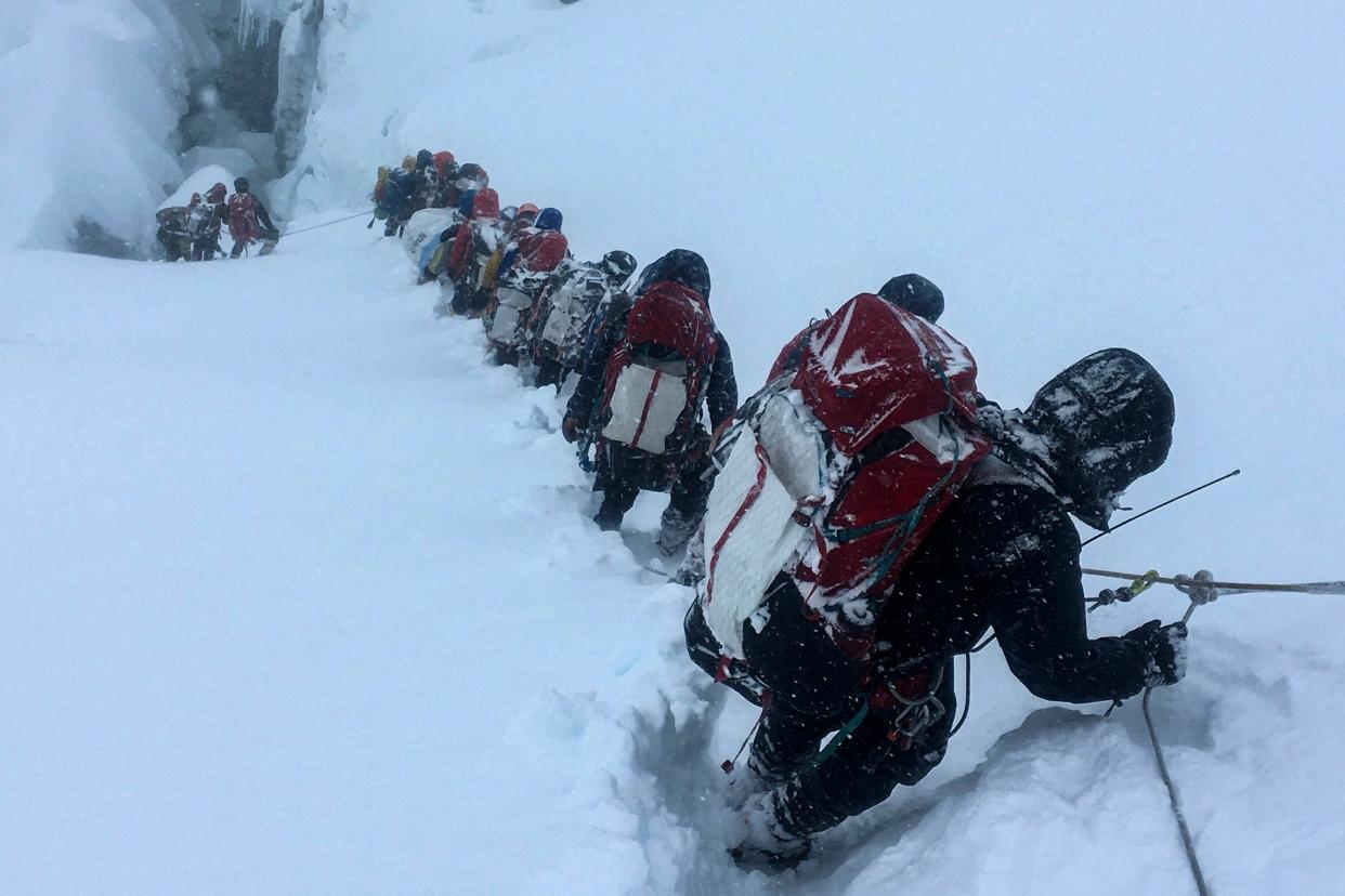 A group of mountaineers who are tethered together by a rope descending a snowy incline while carrying large bags on their backs.