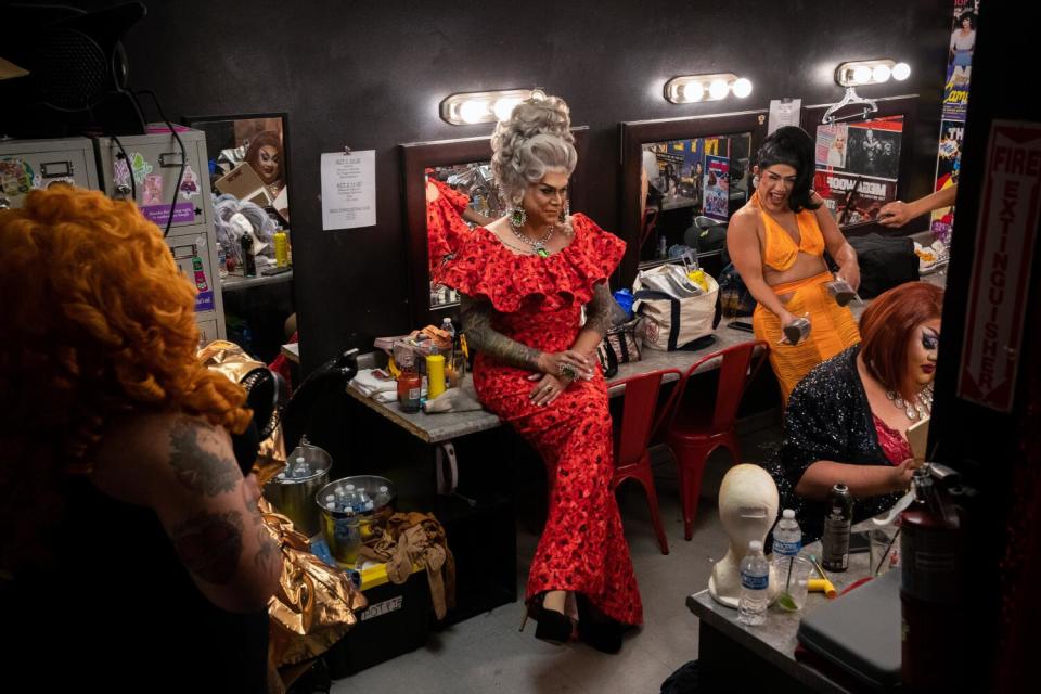 Several drag queens in colorful gowns wait backstage to perform.