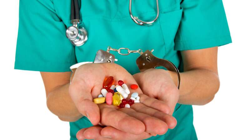 Close-up of a doctor's hands holdings pills, with his wrists handcuffed. Doctor is wearing green scrubs and a stethoscope around his neck.