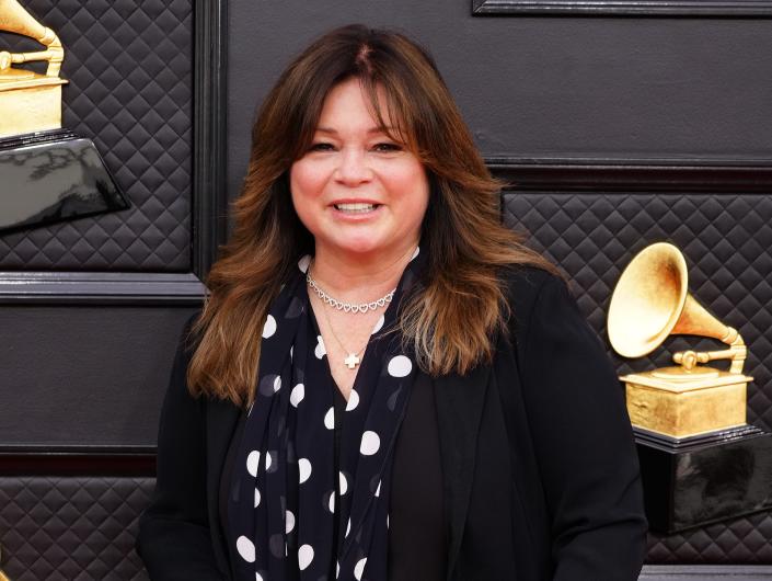 valerie bertinelli in a black and white polka dot shirt and long black cardigan blazer on the grammys red carpet, smiling and holding a clutch that reads &quot;love&quot;