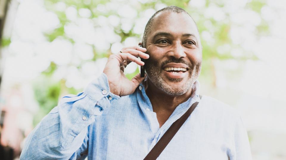 A cheerful African American man in his early 60's smiles, standing outdoors and looking relaxed, confident, and content as he talks on his smart phone.