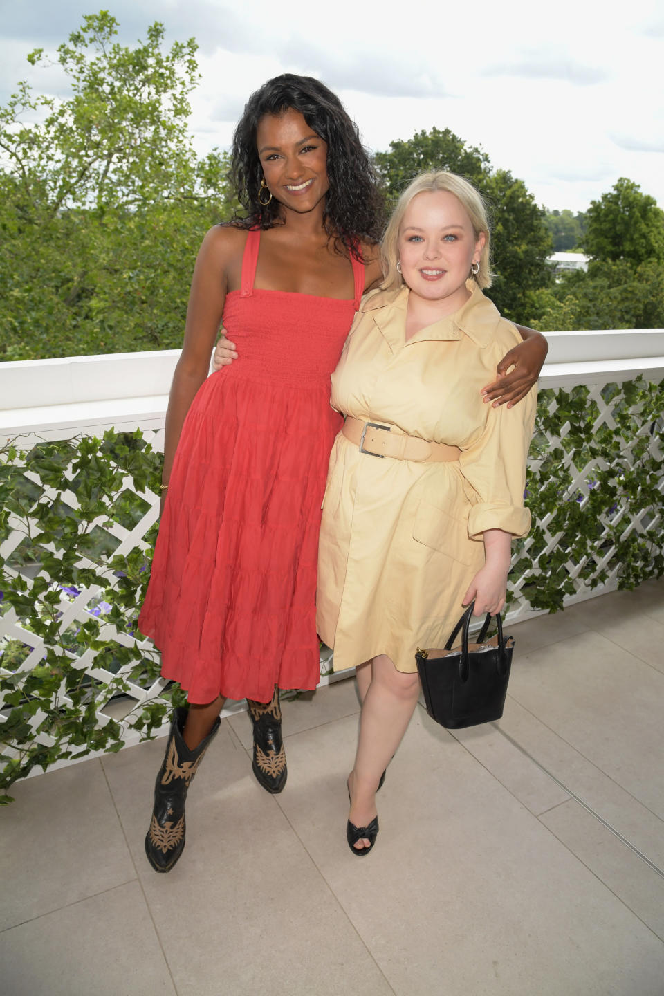 Simone Ashley in a sleeveless dress with cowboy boots stands next to Nicola Coughlan in a belted trench dress holding a black handbag; both are smiling