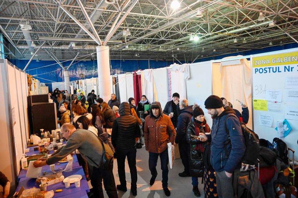 People of all ages, many wearing warm clothes and hats and some with luggage beside them, walk around a long table set out with food.