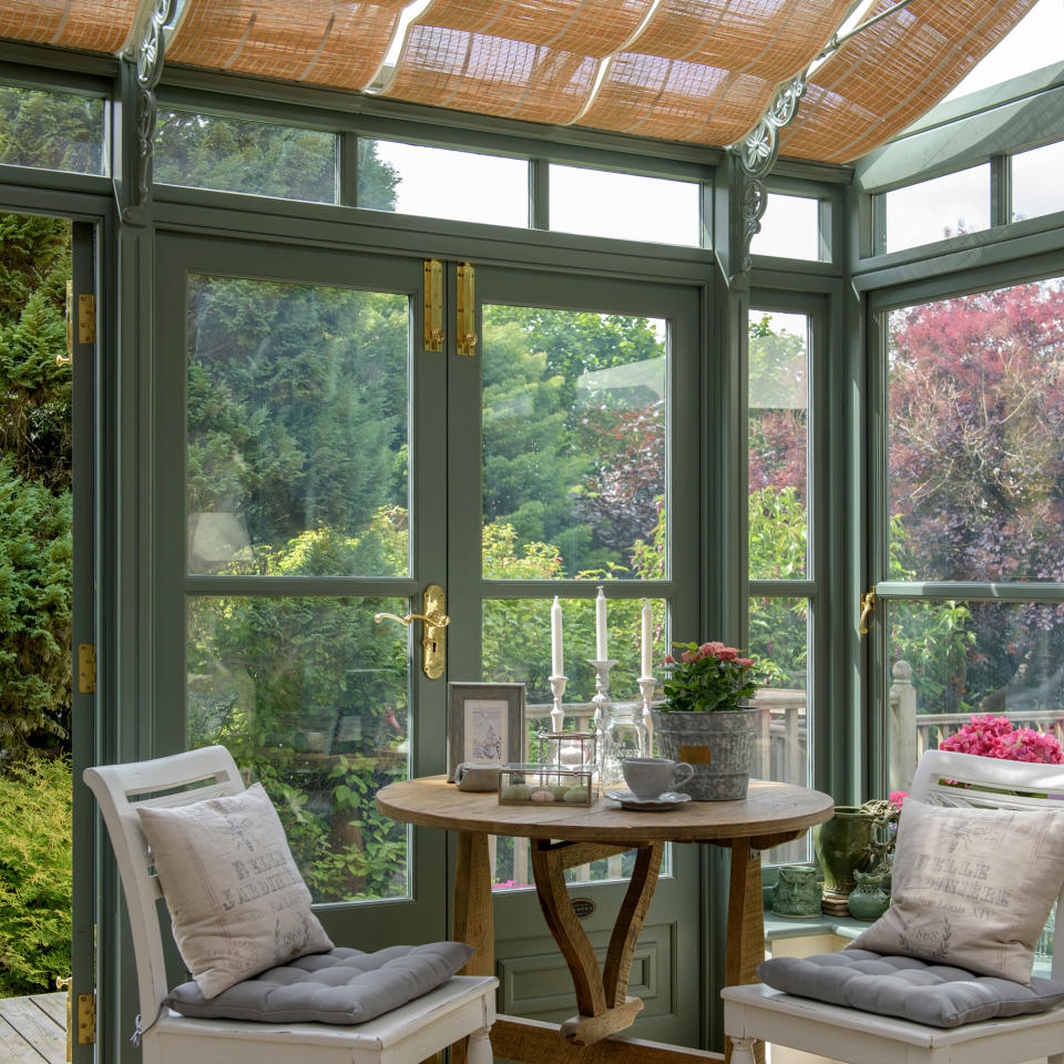 Corner of green-framed conservatory with woven blinds and two dining chairs by round table