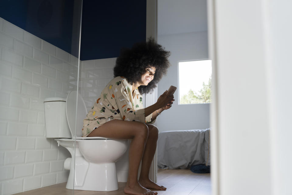 Person sitting on a toilet, smiling while looking at a phone.  They are wearing patterned leisurewear.  The bathroom has a window that provides natural light