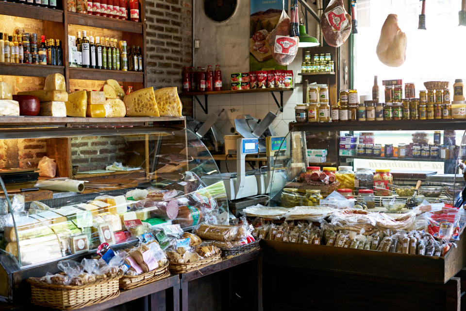 A well-stocked deli with various cheeses, meats, jars of preserves, and packaged foods displayed. The interior suggests a cozy, traditional market ambiance