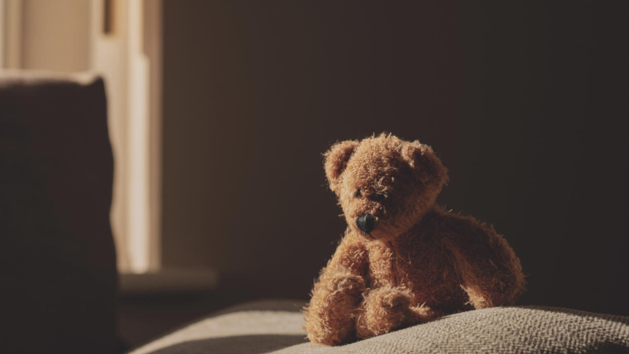  Photo of a teddy bear sitting on a bed in a darkened room with light coming through a window. 