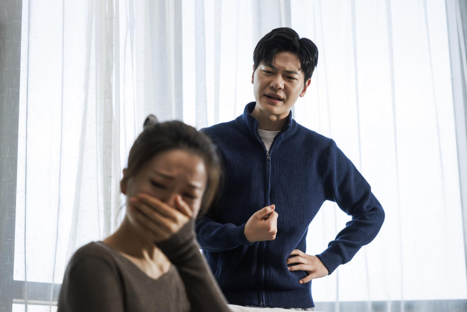 A young Asian couple wearing casual clothes argue in the living room at their home. The man is standing appears angry while the woman is crying and looks sad.