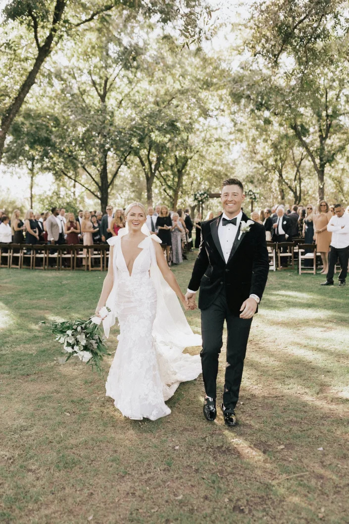 A bride and groom hold hands and exit their wedding ceremony in a wooded area.