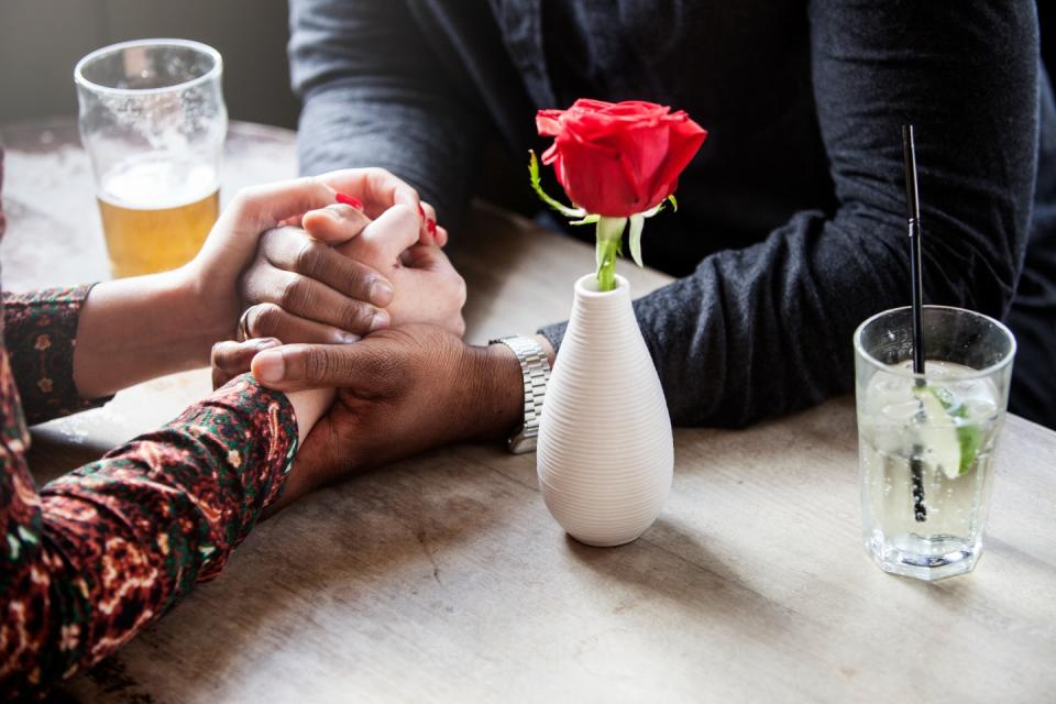 A couple holding hands at a table, with drinks and a rose next to them.