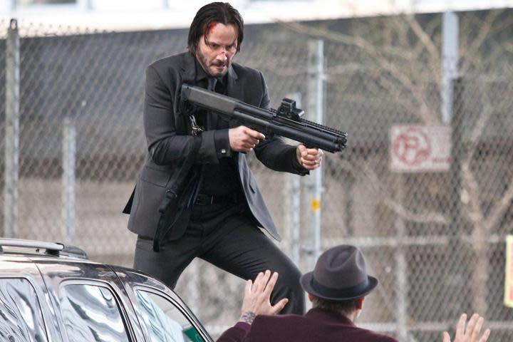 John Wick holds a grenade launcher while standing on a car.