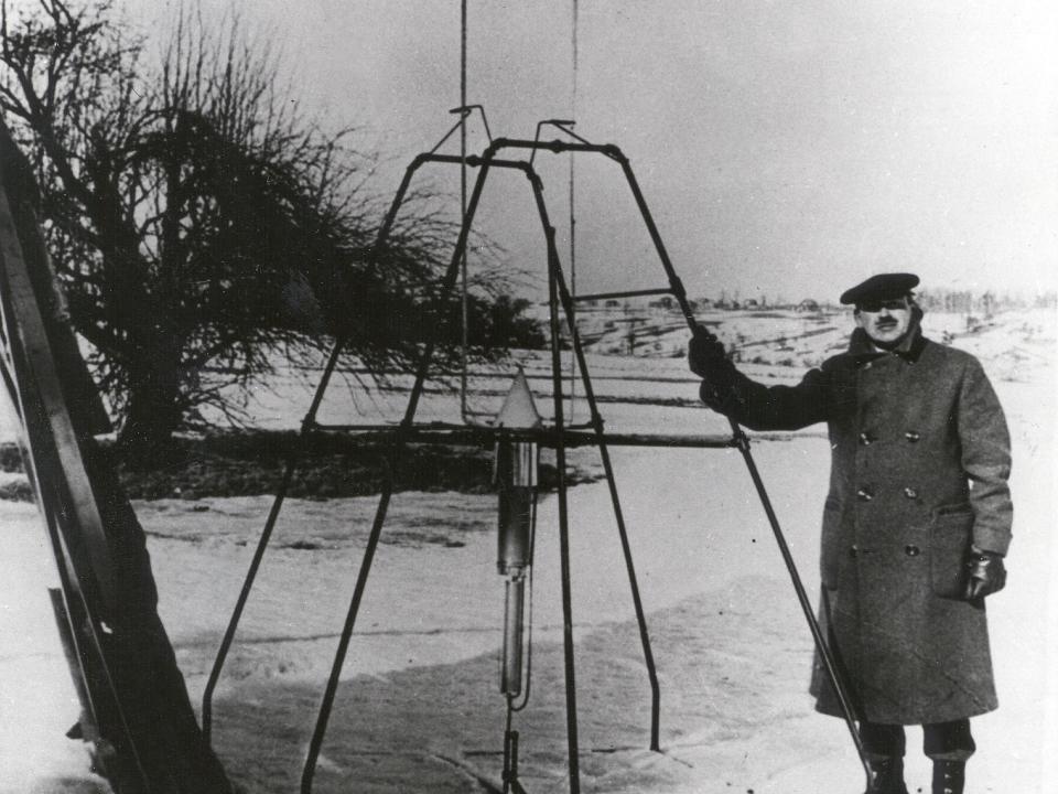 Robert Goddard standing next to a metal pole contraption outside.