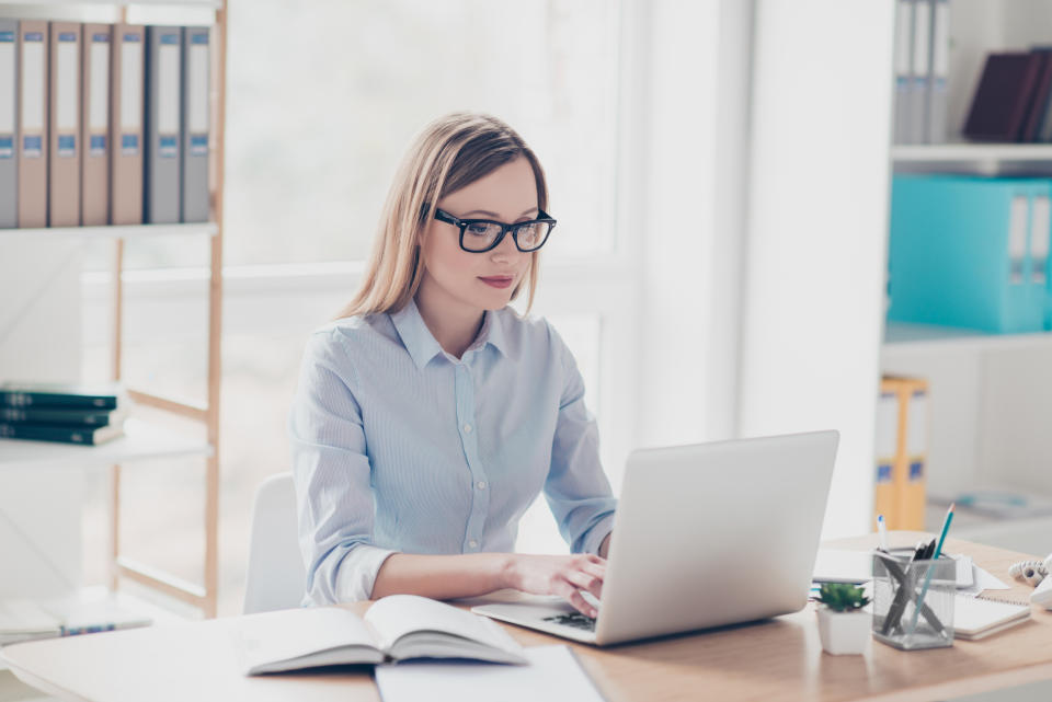 A woman wearing a blue button-down shirt and glasses works on a laptop.