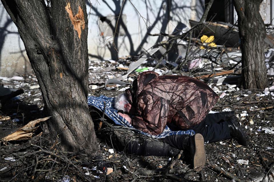 A man with white hair surrounded by debris prostrates himself over a lifeless body lying face down on the ground.