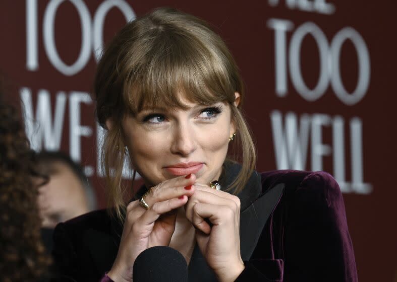 A blond woman with bangs wearing a purple velvet jacket while making a pouting face and cupping her hands under her chin