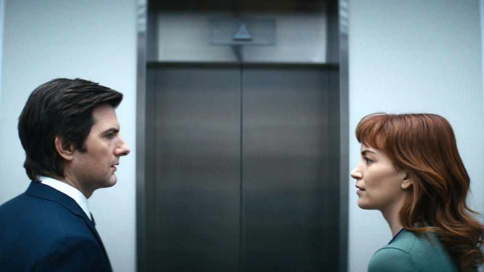 A man and woman face each other in front of an elevator in business clothes. (