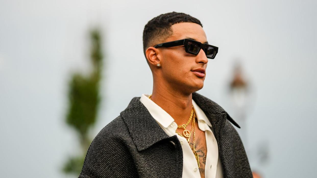 a man wearing sunglasses, stud earrings, layered gold necklaces, a white button down shirt, adn grey coat on the street during paris fashion week