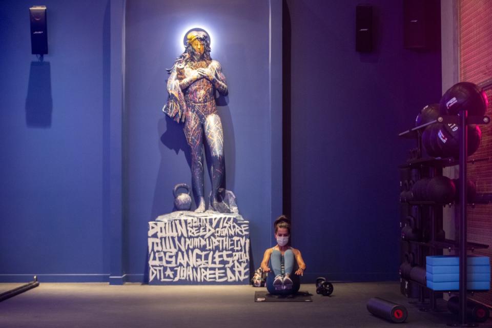 A woman sits on a mat to do exercises next to a statue with a kettlebell at its feet