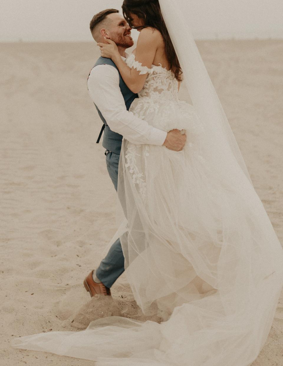 A groom lifts his bride up on a beach and they lean their heads together.