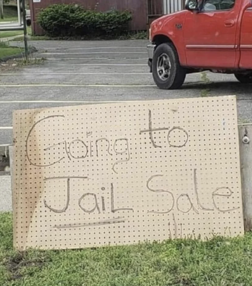 Handwritten sign reading "Going to Jail Sale" placed on grass near a sidewalk and vehicle