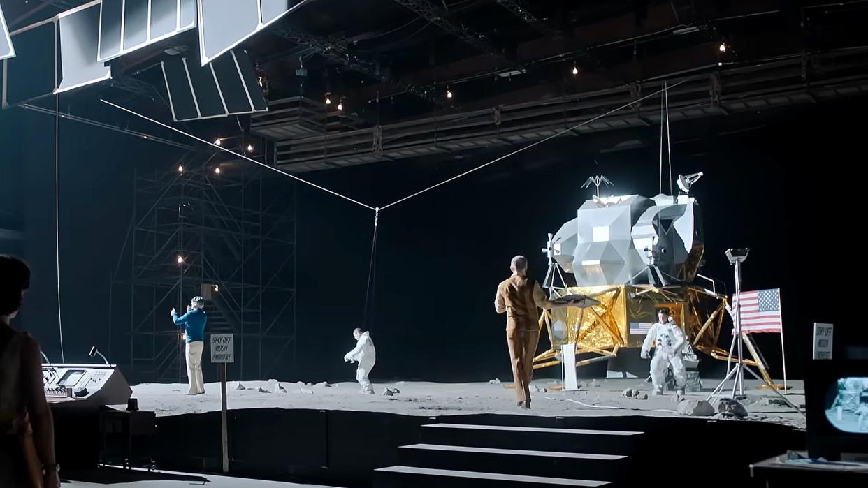  People in white spacesuits walk across a movie set built to resemble the surface of the moon. 