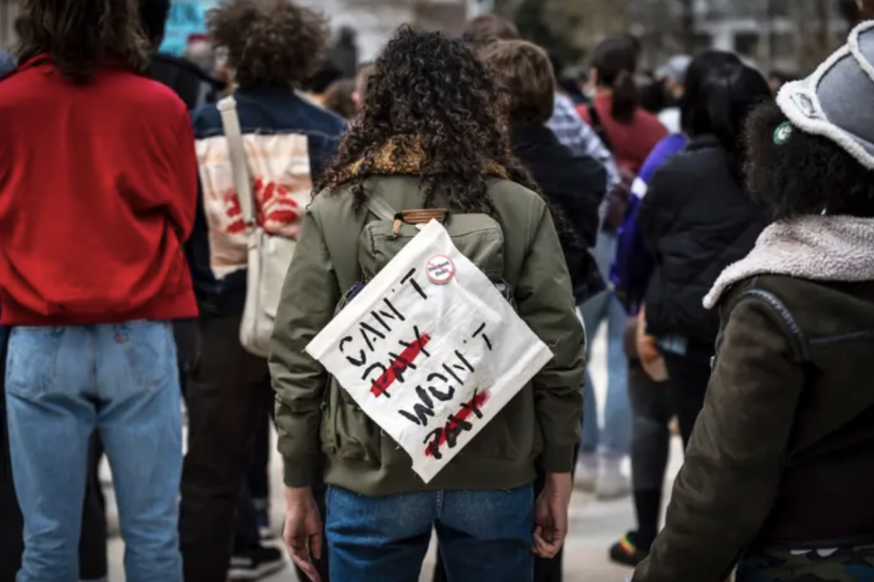 A person in a green jacket and curly hair stands in a crowd with their back to the camera. on their back is a canvas sign that says "can't pay, won't pay."