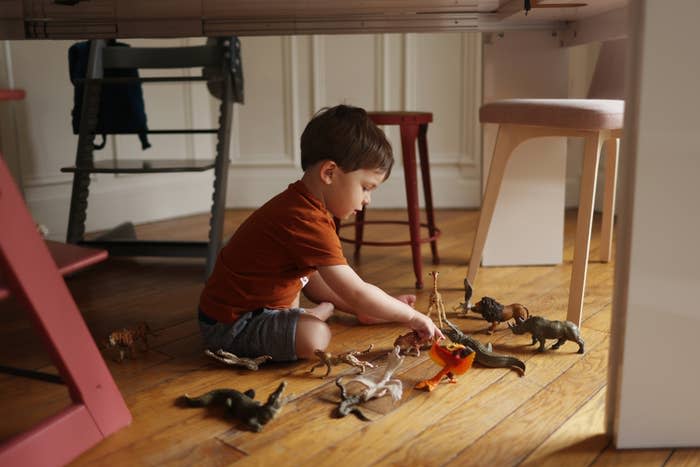 A young child in a red shirt and shorts sits on the floor playing with toy dinosaurs and animals under a table