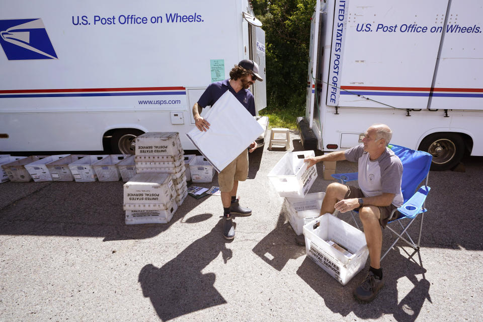 Postal Clerk Randy Boucher, right, hands mail to Wyatt Galfetti at a makeshift post office created from two trucks in a college parking lot, Tuesday, Aug. 1, 2023, in Montpelier, Vt. The mostly gutted shops, restaurants and businesses that lend downtown Montpelier its charm are considering where and how to rebuild in an era when extreme weather is occurring more often. Vermont's flooding was just one of several major flood events around the globe this summer that scientists have said are becoming more likely due to climate change. (AP Photo/Charles Krupa)