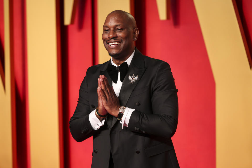 Tyrese Gibson smiling, wearing a black tuxedo with a brooch pin and bow tie, hands clasped together, standing against a red backdrop with large letters