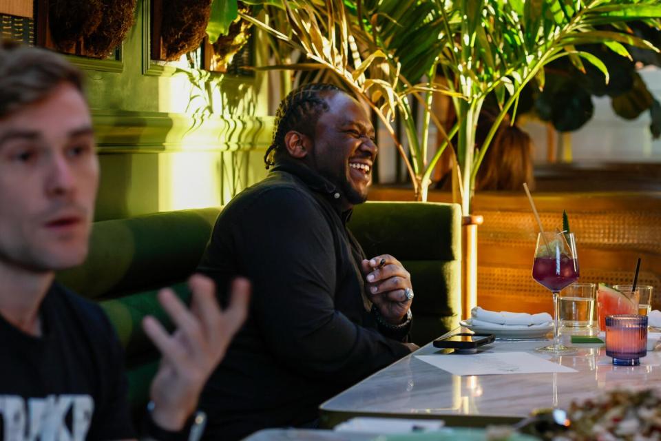 A man in a restaurant laughs while a man in the foreground gestures.