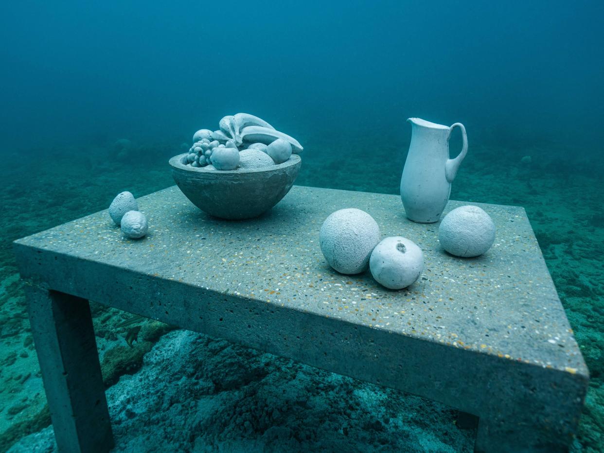 An underwater sculpture of a table with a bowl of fruit, a pitcher, and loose fruit.