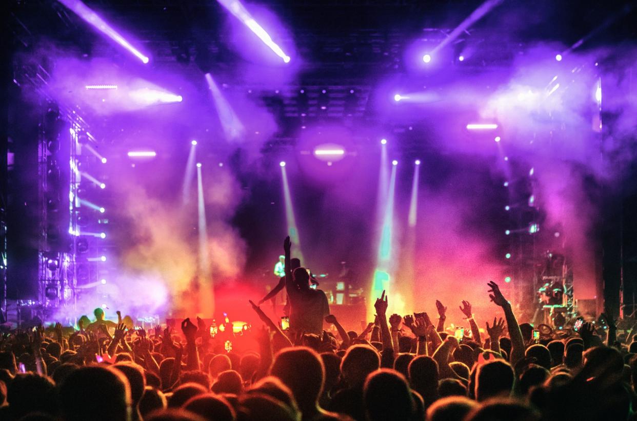 Rear view of large group of people enjoying a concert performance. There are many hands applauding and taping the show. Blue and red spot lights firing from the stage.Each individual inspected for recognizability.