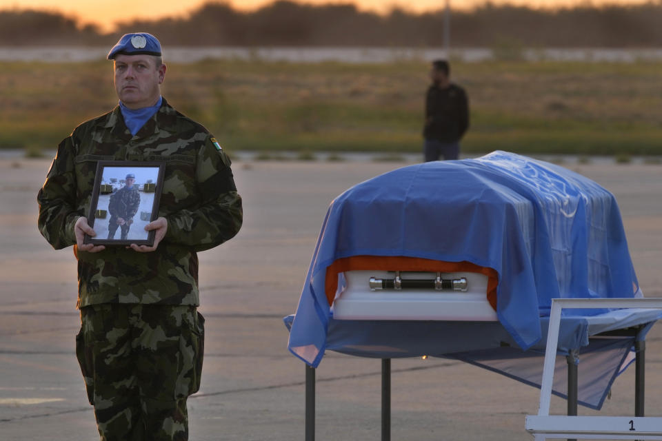 An Irish U.N peacekeeper, stands next to the coffin draped by the United Nations flag of his comrade Pvt. Sean Rooney who was killed during a confrontation with residents near the southern town of Al-Aqbiya on Wednesday night, as he holds his portrait during his memorial procession at the Lebanese army airbase, at Beirut airport, Sunday, Dec. 18, 2022. (AP Photo/Hussein Malla)