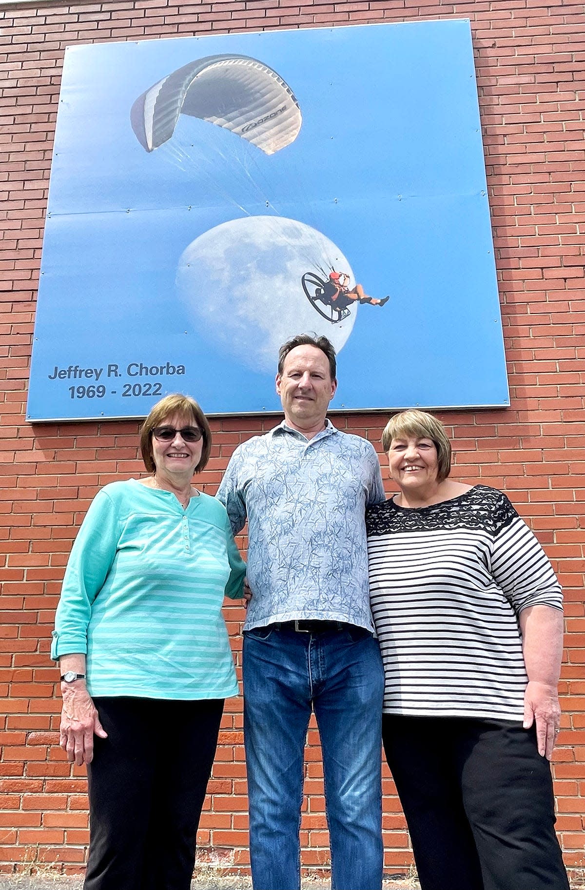 Joe Parker (center) is joined by Eileen Chorba (left) and Kathy Taninies for the unveiling of a tribute to Jeff Chorba, who tragically died in a paramotoring accident one year ago. The 8x8 image will be on display all through the summer on the exterior of the Prompton Tool building, located at Sunrise Avenue in Honesdale.