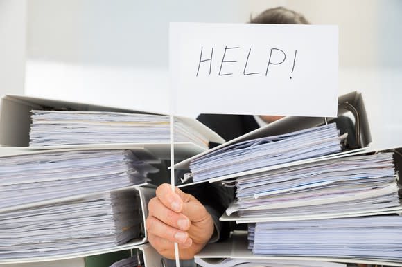 A hand with a small help! sign at a desk buried in paperwork.