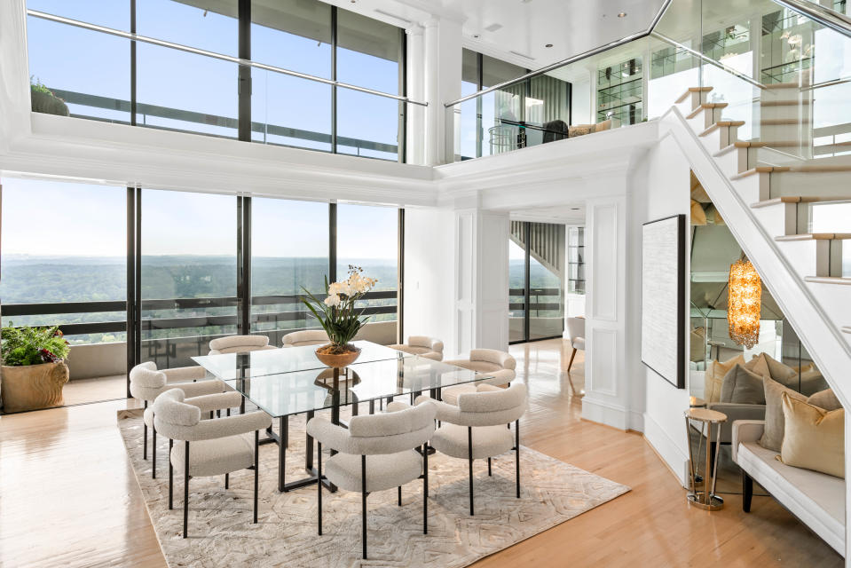 A living room with boucle arm chairs and large floor to ceiling windows and a mirror in the nook under the stairs