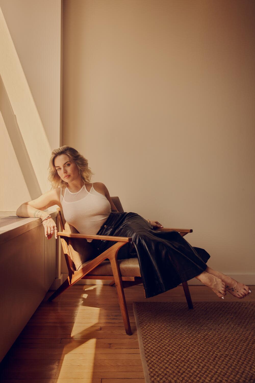 A blond woman in a white tank top and black leather skirt sits on a chair with feet stretched.