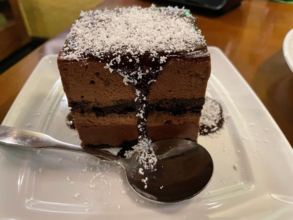 Slice of chocolate cake with chocolate sauce dripping from top to a spoon on the plate. The cake has distinctive chocolate layers